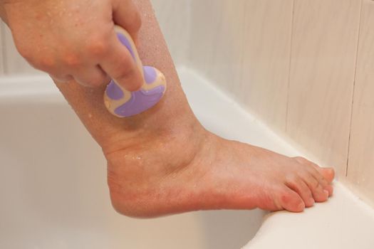 Close up of a razor shaving hair from a leg on a tub or shower stall