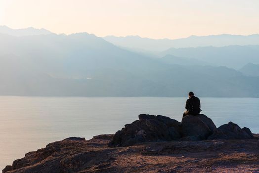 A man sits on a high mountain cliff and overlooks the horizon. A concept for hope, peace, power, soul searching. Desert mountains against the backdrop of the Red Sea. Shlomo mountain, Eilat Israel