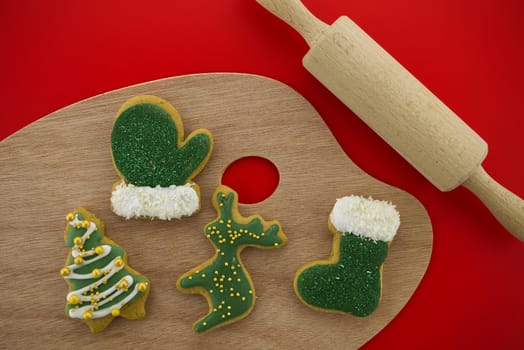 Christmas food and baking concept with a selection of colorful glazed or iced cookies in different shapes on a board with rolling pen over a festive red background in a flat lay still life