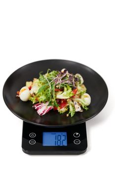 Close up of appetizing vegetables salad on black kitchen scales on white background. Concept of proper and balanced nutrition and weight of food. 