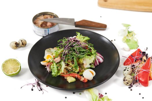 Close up of modern black plate with fresh greens,eggs,peppers and tuna in its own juice on white background. Concept of preparing cooking vegetables salad with fish.