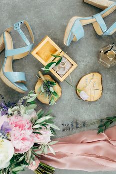 Wedding rings lie in a wooden box surrounded by sandals with heels, flowers and twigs, a bouquet and a scarf on the floor. High quality photo