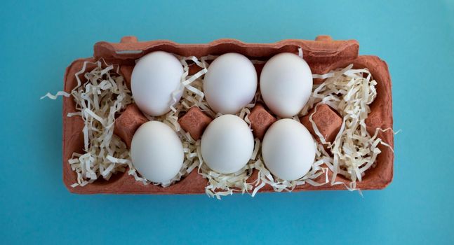 Six white fresh organic eggs in a beige eco-friendly cardboard egg container on a blue background. Easter concept, ecology, zero waste.