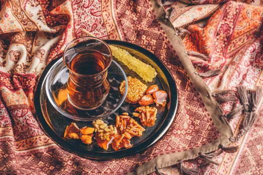 Black tea in armudu glass with candies with different nuts and crystal sugar on metal tray over tablecloth