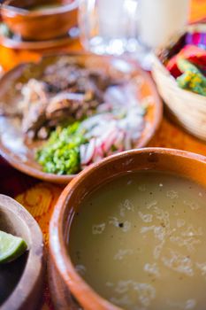 Close-up of clay bowl of lamb broth and plate of chopped lamb meat above orange and red tablecloth. Traditional soupy dish and condiments above colorful table. Classic Mexican cuisine