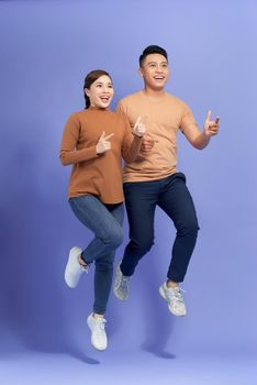 Excited young Asian couple man and woman jumping and pointing with happy and fun over purple background