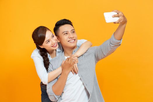 Young and beautiful couple expressing joy on camera while partying and taking selfie isolated over yellow background.