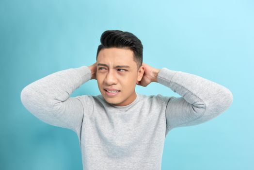 Emotional stress. Frustrated young man holding hands on ears while standing against blue background