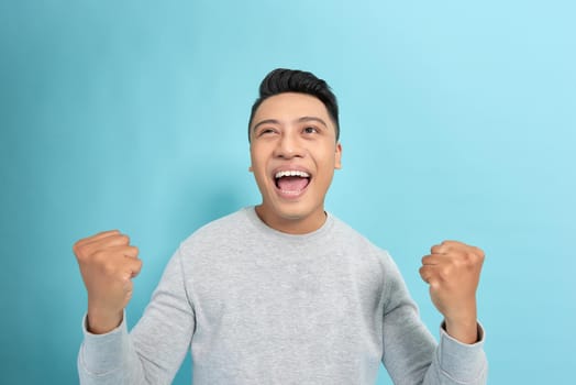 Overjoyed male clenches fists with happiness, opens mouth widely as shouts loudly, celebrates his success, poses against blue background. 