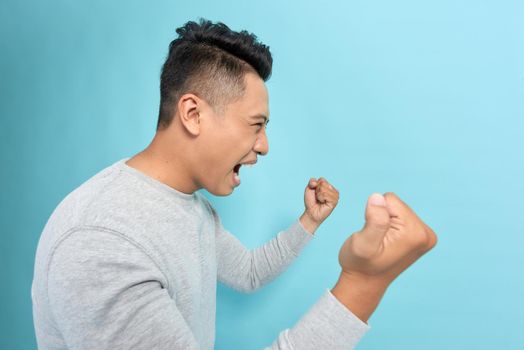 Overjoyed male clenches fists with happiness, opens mouth widely as shouts loudly, celebrates his success, poses against blue background. 