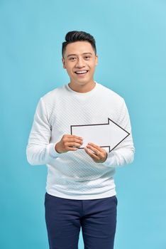 Happy young businessman holding a arrow pointing right and leaning against a wall isolated on blue background