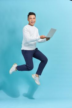 Image of young man dressed standing over blue background using laptop computer while jumping.