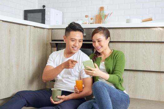 Laughing guy with a girl sitting on the floor in the kitchen with cups of coffee and orange juice
