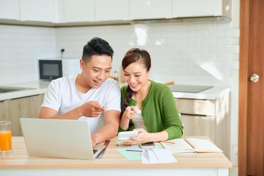 Photo of cheerful loving young couple using laptop and analyzing their finances with documents. Look at papers.