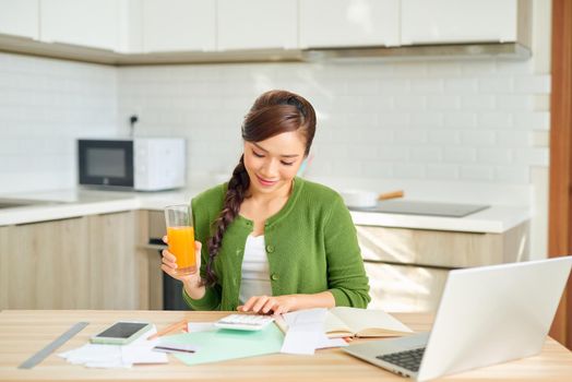 Attractive young smiling Woman paying bills online using laptop from home while sitting in the kitchen