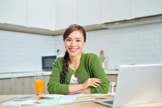 Smiling young woman sit at desk in kitchen working on laptop gadget, happy millennial girl manage finances pay household expenditures or taxes online,