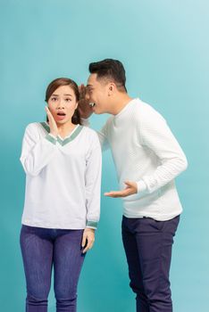 Asian man sharing secret or whispering gossips into his girlfriend's ear, blue studio background
