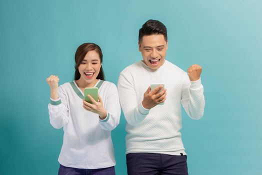 Surprised young couple stand isolated on blue background hold smartphones happy win online lottery,