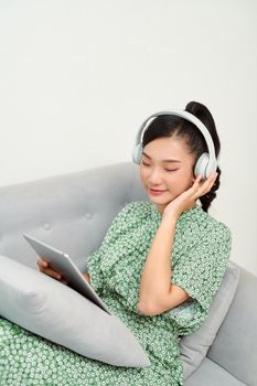 Young beautiful Asian woman laying in sofa and playing tablet with headphones 