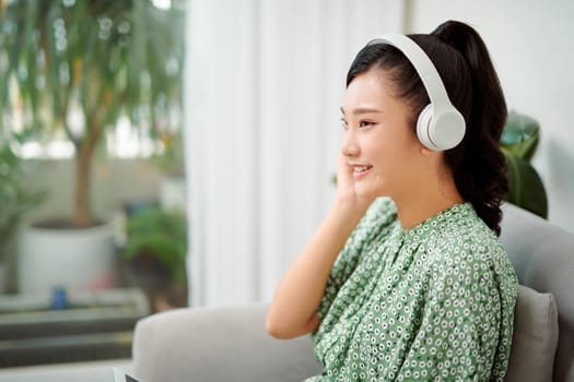 Happy woman wearing wireless headphones listening to music sitting on a couch in the living room at home