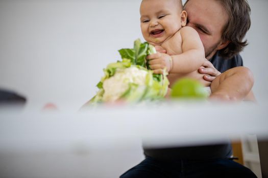 Father lovingly holding and kissing his happy baby daughter above table. Adorable baby smiling and playing with cauliflower. Babies interacting with food