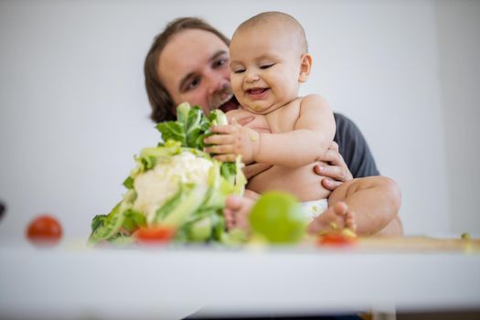 Father lovingly holding and kissing his happy daughter above table with vegetables. Adorable smiling baby on wooden board playing with cauliflower. Babies interacting with food