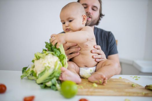 Father lovingly holding his happy baby daughter above table with vegetables. Adorable baby on wooden board playing with cauliflower. Babies interacting with food
