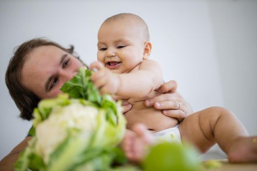 Father lovingly holding and kissing his happy baby daughter above table. Adorable baby smiling and playing with cauliflower. Babies interacting with food