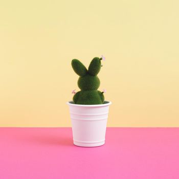 Green easter bunny decorated with flowers in flowerpot on vibrant yellow and pink background. Minimalist concept. Square with copy space.