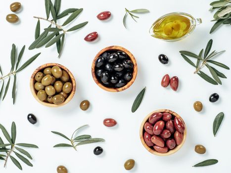 Set of green olives, black olives and red kalmata olives and extra virgin olive oil on white background. Top view of different types of olives in bowls and oil on leaves and branches background.