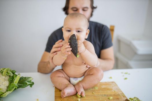 Father lovingly holding his happy baby daughter above table. Adorable baby sitting on wooden board and biting avocado peel. Babies interacting with food