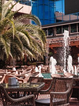 Empty tables in cafe or restaurant on open air, with palm tree and fountain. Street cafe interior. Vacation and resort concept. Vertical.