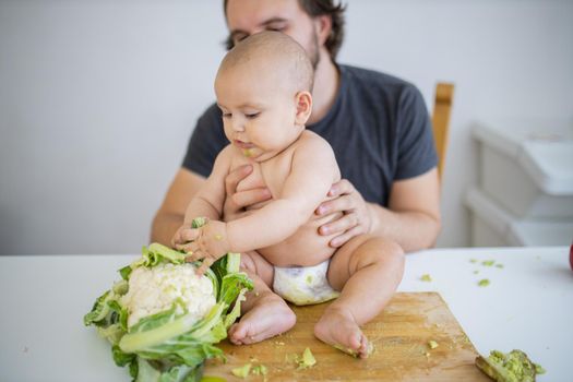 Father lovingly holding his happy baby daughter above table. Adorable baby on wooden board playing with cauliflower. Babies interacting with food