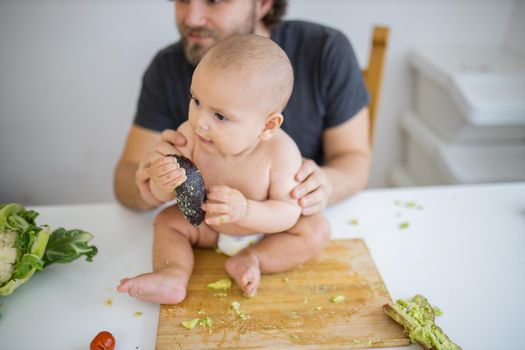 Father lovingly holding his happy baby daughter above table. Adorable baby sitting on wooden board and holding avocado peel. Babies interacting with food