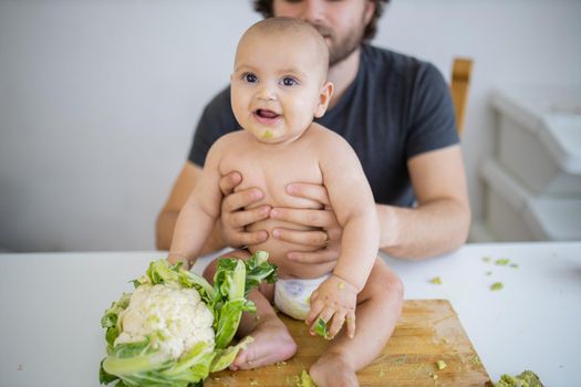 Father lovingly holding his happy baby daughter above table. Adorable baby on wooden board smiling and holding cauliflower. Babies interacting with food