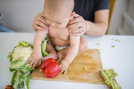 Father lovingly holding his happy baby daughter above table. Adorable baby on wooden board playing with bell pepper. Babies interacting with food