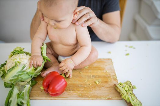 Father lovingly holding his happy baby daughter above table. Adorable baby on wooden board playing with cauliflower. Babies interacting with food