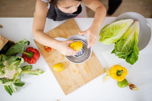 Adorable little girl squeezing lemon on steel lemon juicer surrounded by vegetables. Young child making juice on white table from above. Kids preparing food