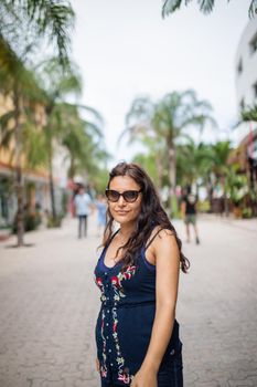 Beautiful woman wearing blue dress standing in a tropical street with palm trees as background. Portrait of smiling woman wearing sunglasses with blurry background. Tropical summer vacations