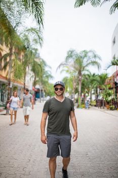 Bearded mas wearing sunglasses and hat walking in a tropical street with palm trees as background. Portrait of handsome man with blurry background. Tropical summer vacations