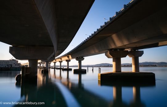 Sweeping structural lines of Tauranga Harbour Bridge from below over calm blue water. Bridge crosses Tauranga harbour to Mount Maunganui.