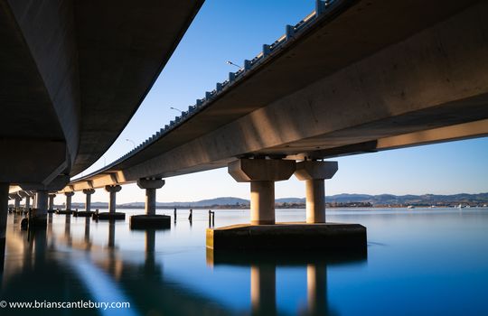 Sweeping structural lines of Tauranga Harbour Bridge from below over calm blue water. Bridge crosses Tauranga harbour to Mount Maunganui.