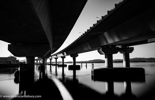 Sweeping structural lines of Tauranga Harbour Bridge from below over calm water in monochrome. Bridge crosses Tauranga harbour to Mount Maunganui.