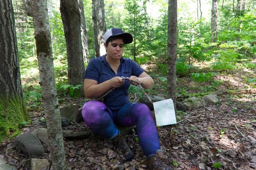 Woman focused on using a knife in the woods to cut kindling for firewood