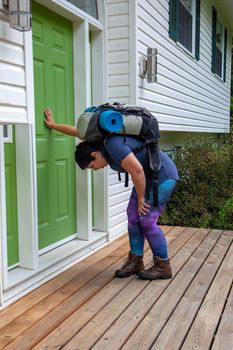 person leaning against their front door with backpack and camping roll after a trip 