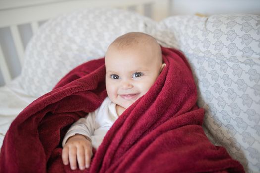 Adorable and happy baby covered with red blanket and lying on bed. Cute baby smiling and resting on pillows. Toddles and babies on beds