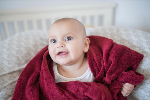 Adorable and happy baby covered with red blanket and lying on bed. Cute baby smiling and resting on pillows. Toddles and babies on beds