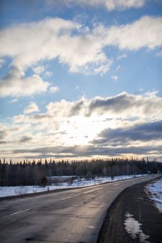 A road in the background of nature. Open road in the future, no cars, cars on the paved road through the green forest, trees. Clouds and the sun in the blue sky in the spring, in the evening. 