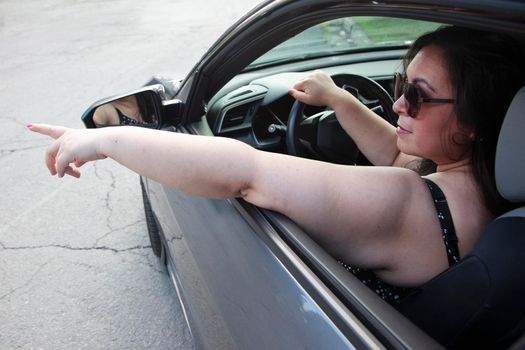 woman with sunglasses points out the window while driving her car