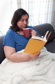 Real latina woman cooling off her mug of hot drink while reading a hardcover novel at home